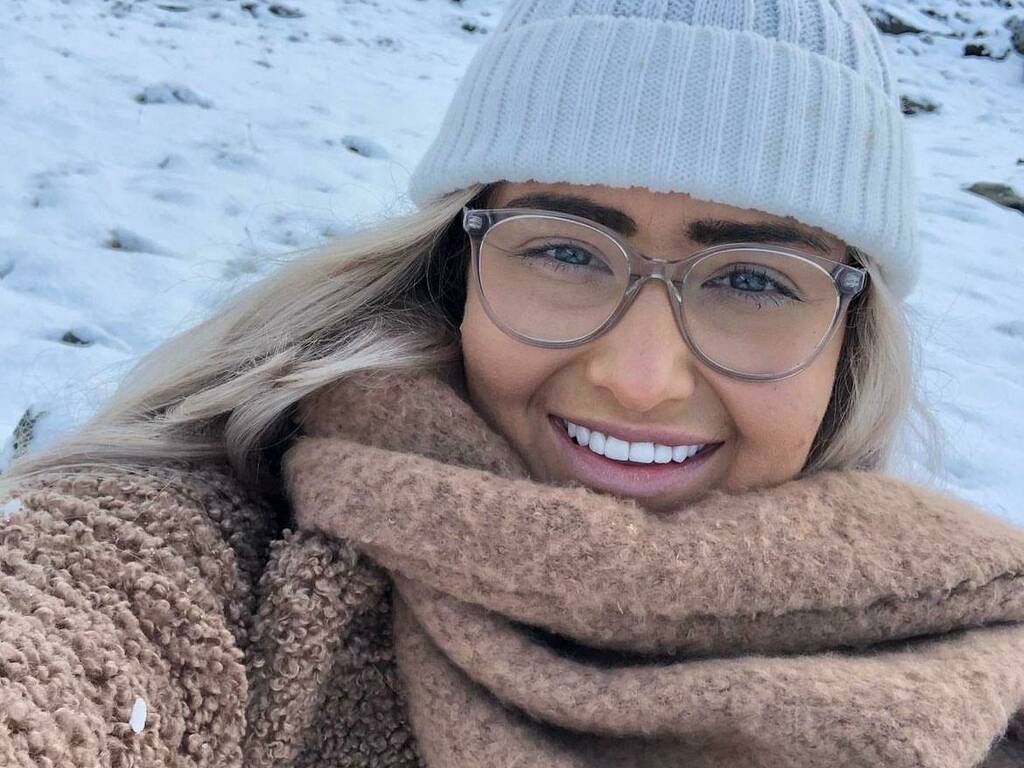 Jasmine smiling wearing a white woollen hat, beige coat and scarf with snow in background.